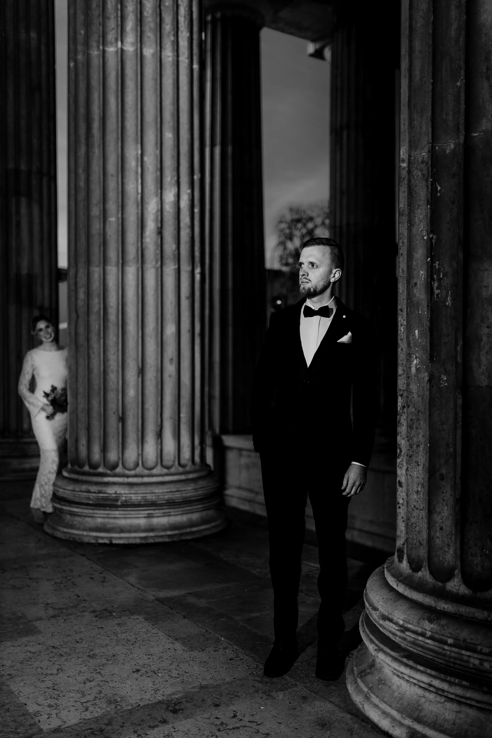 Man in tuxedo between the columns of Königsplatz Munich in black and white – couple photoshoot